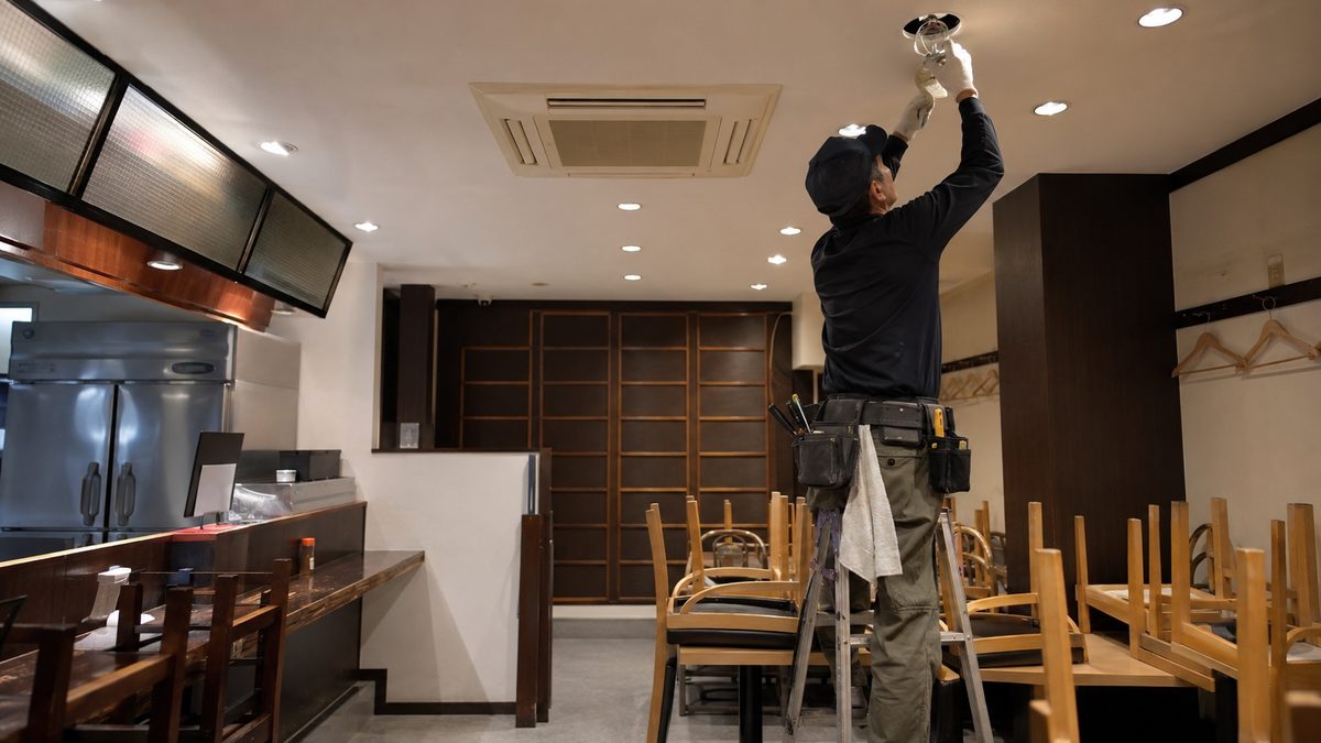 小さな店舗で脚立に立ち天井照明を扱う職人（AI・構成見本）